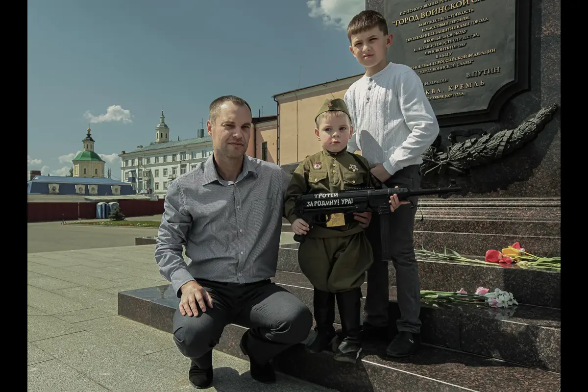 A family of collectors of Soviet memorabilia with a six-year-old boy dressed in a 1940s Soviet military uniform. Inscription on the wooden rifle: “Trophy for the Motherland! Hurrah!” In the background, a stele bearing Putin’s words awarding Yelets the title of “City of Military Glory.”